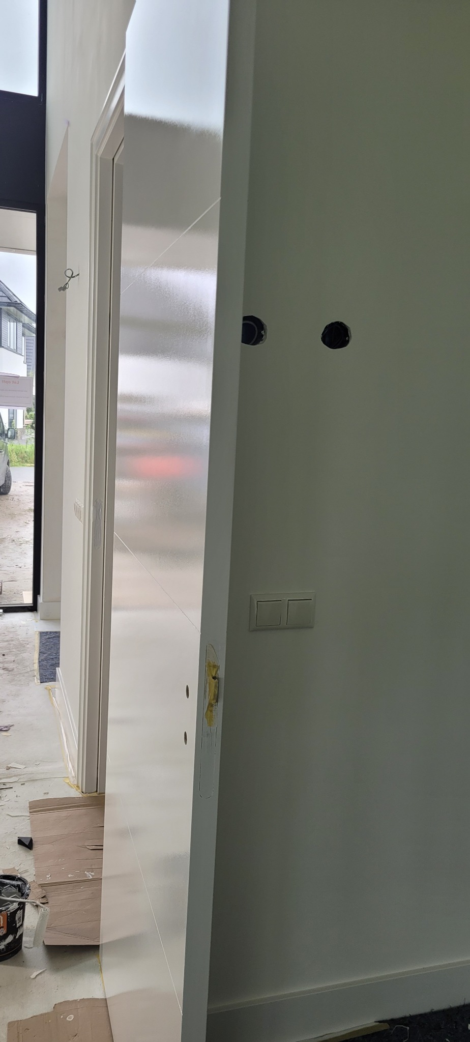 Świeżo pomalowane, białe wnętrze z widocznymi puszkami elektrycznymi i włącznikiem światła. Otwarta, pomalowana na biało ościeżnica drzwiowa z widocznymi śladami taśmy malarskiej zabezpieczającej...