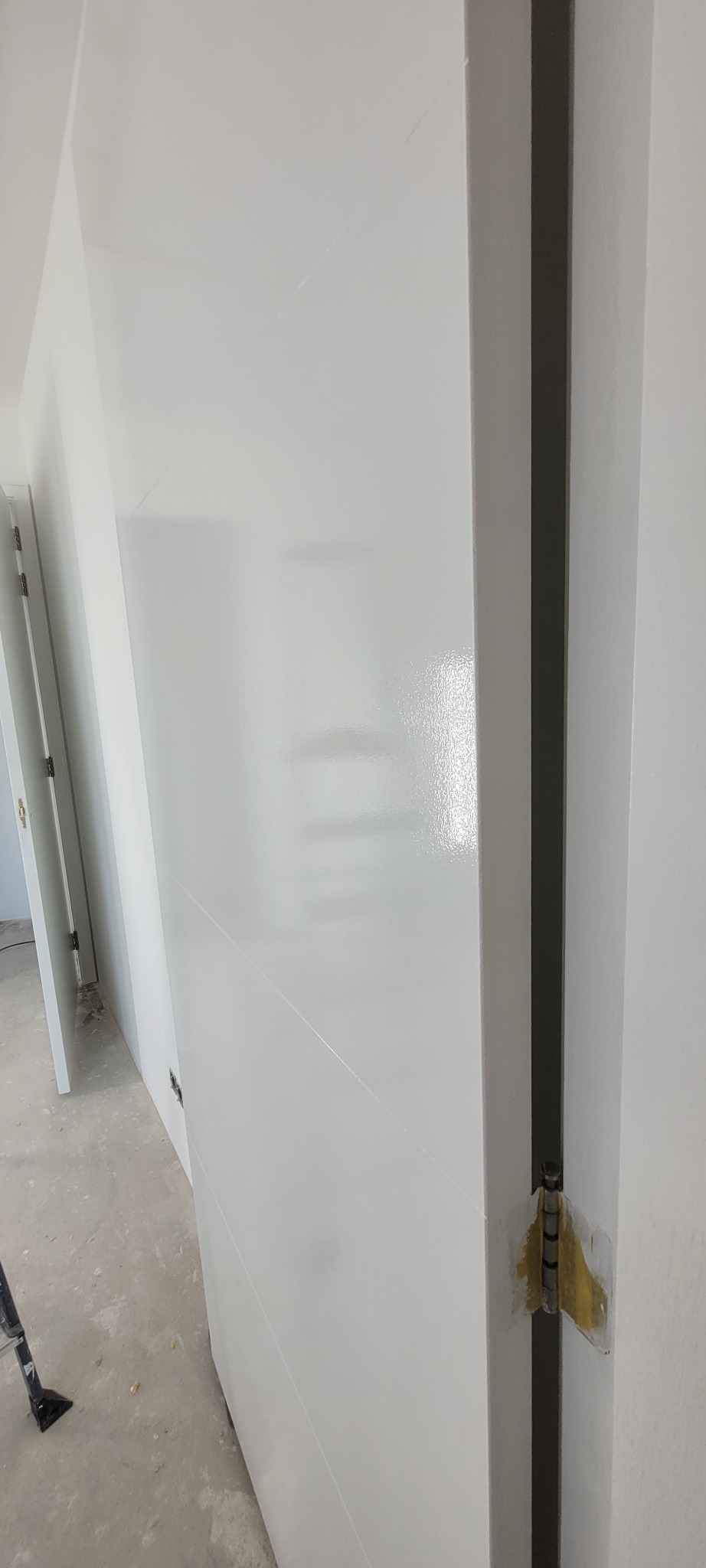 Świeżo pomalowane na biało drzwi wewnętrzne z widocznymi zawiasami zabezpieczonymi taśmą malarską, w tle fragment ściany i podłogi w trakcie remontu.