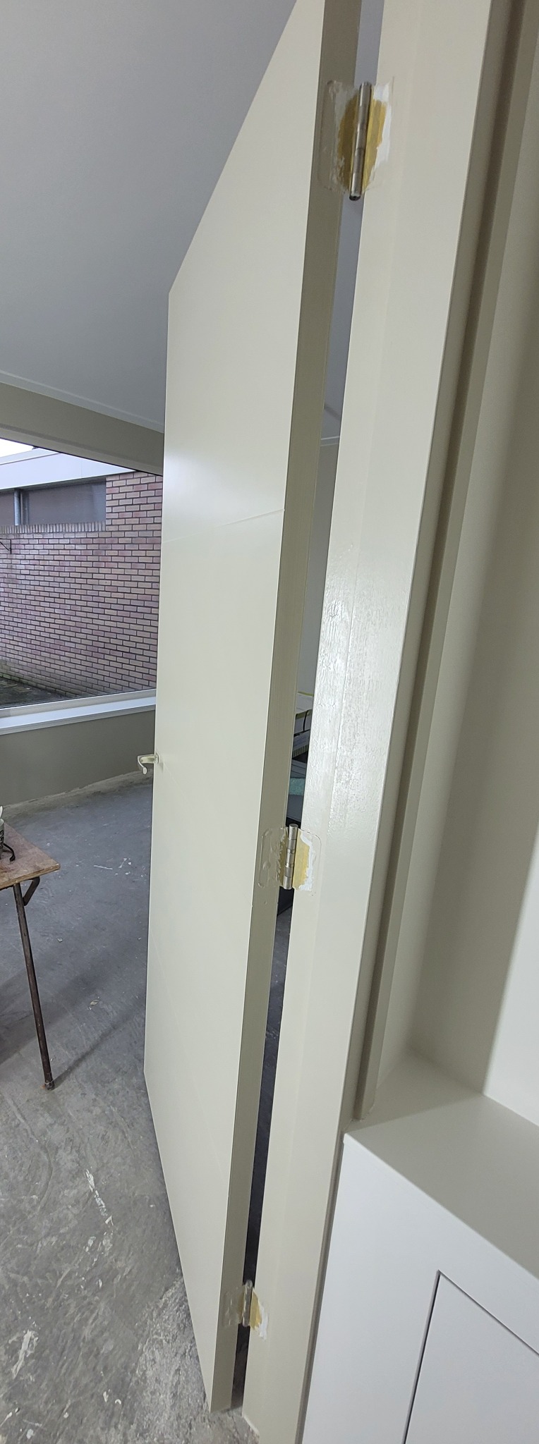 Otwarta, świeżo pomalowana na kremowo rama drzwi z zawiasami zabezpieczonymi taśmą malarską, widok na zewnętrzny mur z cegły.