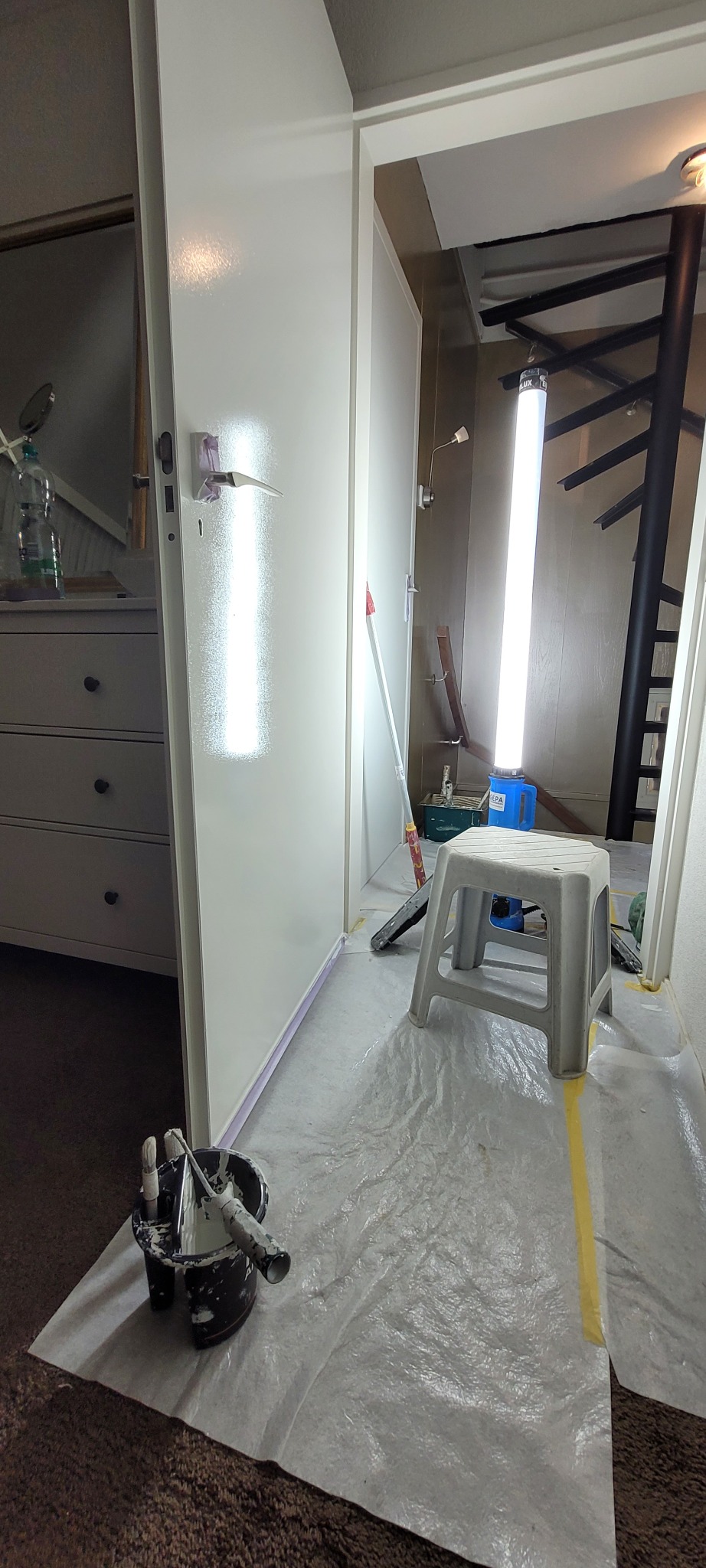Pokój w trakcie odświeżania: świeżo pomalowana na biało ściana z widocznym odbiciem światła, podłoga zabezpieczona folią malarską, wiadro z farbą i narzędziami malarskimi, drabina i lampa...