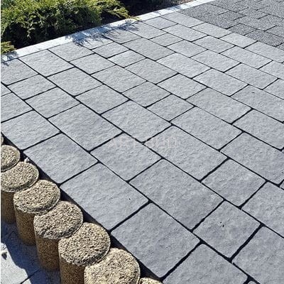 Szare, prostokątne kostki brukowe ułożone pod kątem. Obrzeże z okrągłych, kamiennych elementów. W tle fragment nawierzchni z drobnego kruszywa.