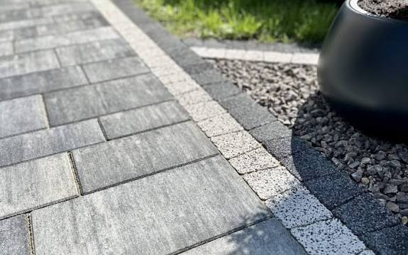 Szara kostka brukowa ułożona wzdłuż ścieżki z obrzeżami z jaśniejszej kostki i czarnym kamieniem dekoracyjnym, obok czarna donica z ziemią i trawa.