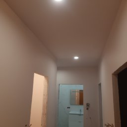 Igor Kaliuta - Korytarz po malowaniu, widoczne gładkie, jasne ściany i sufit z zamontowanymi lampami. W tle wykończona łazienka z umywalką i lustrem. Ościeżnice drzwiowe surowe.