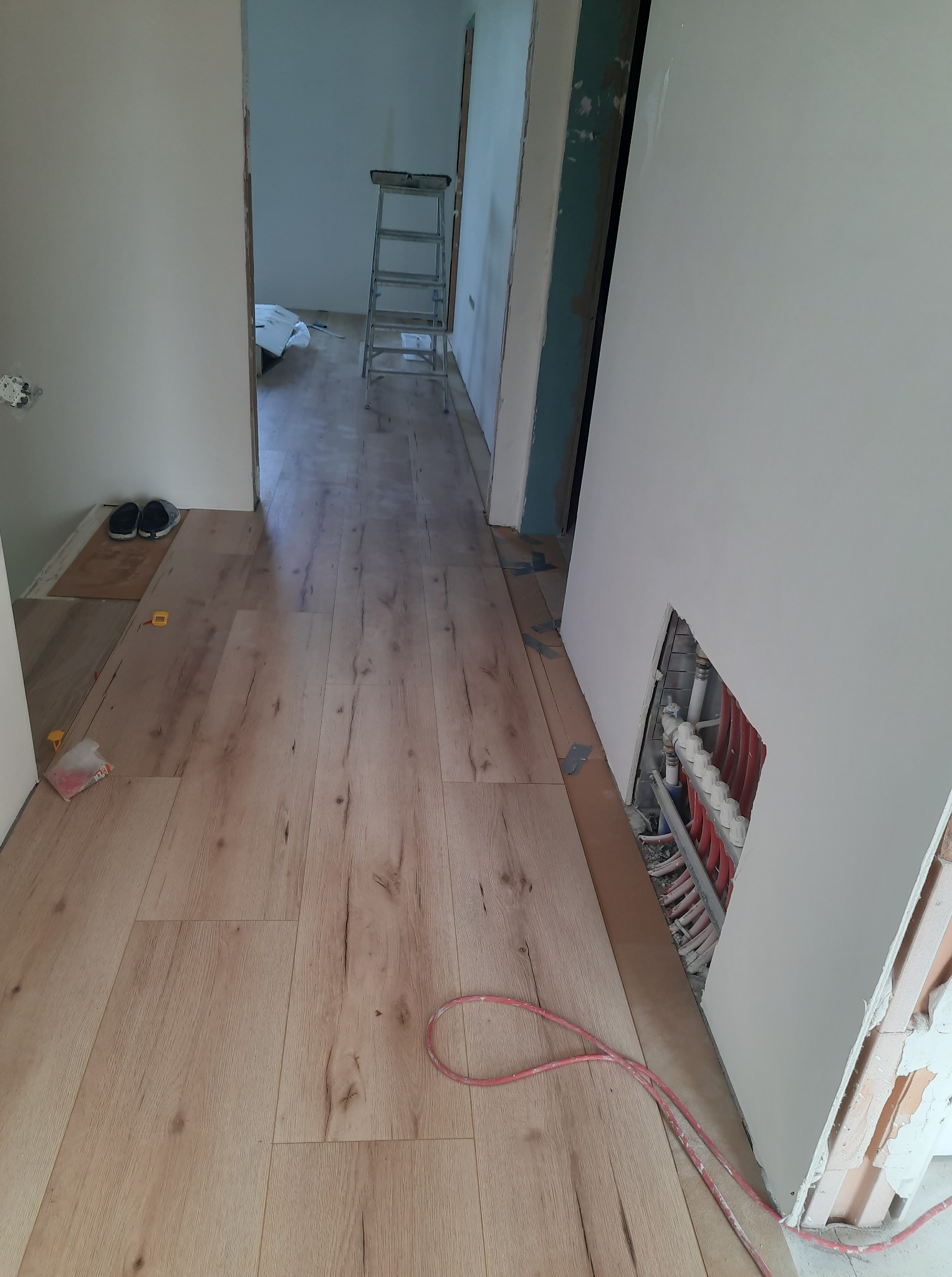 Korytarz w trakcie remontu: ułożone panele podłogowe, ściany przygotowane do malowania, widoczne rury ogrzewania podłogowego w ścianie, drabina w tle.