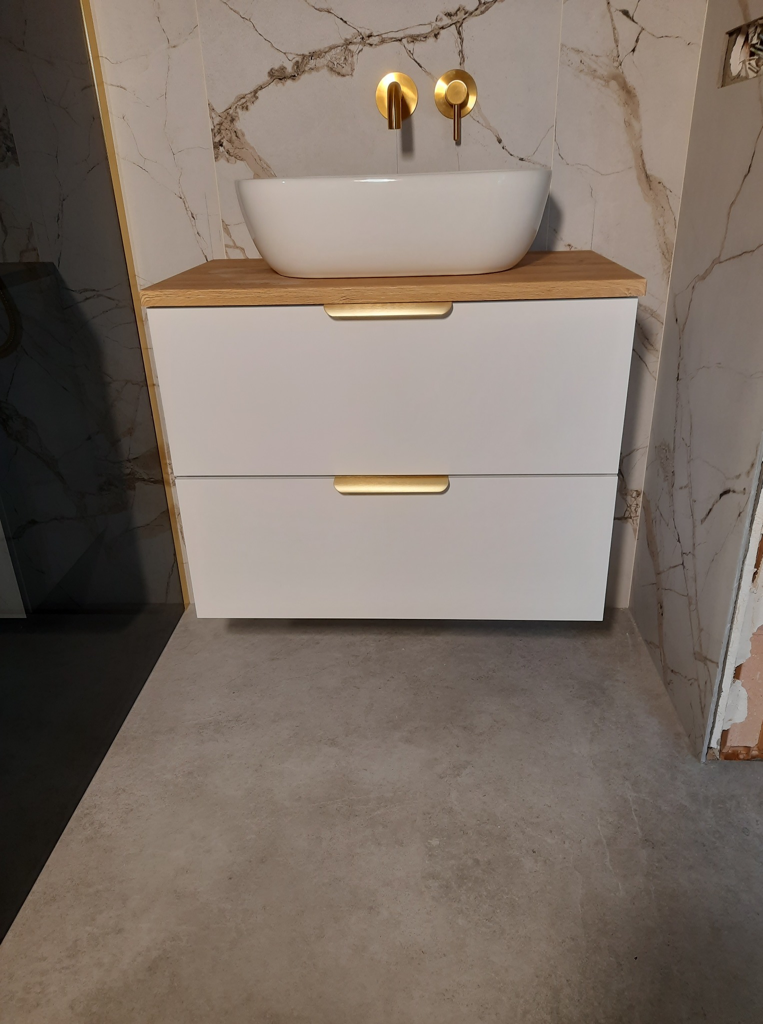 Nowoczesna łazienka z białą szafką pod umywalkę, drewnianym blatem i złotymi akcentami na tle marmurowej ściany. Widoczne złote baterie i szara podłoga.