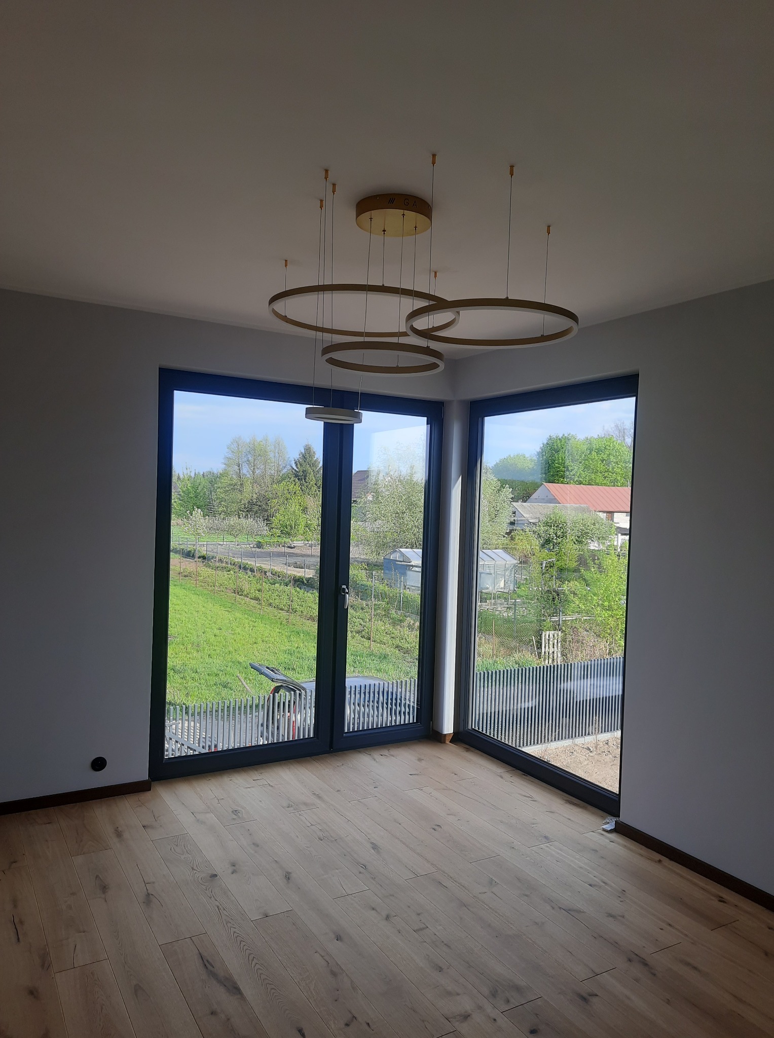 Jasne, puste wnętrze z parkietem i dużymi oknami z widokiem na zieleń. Minimalistyczny, okrągły żyrandol w kolorze złota. Świeżo pomalowane, szare ściany.