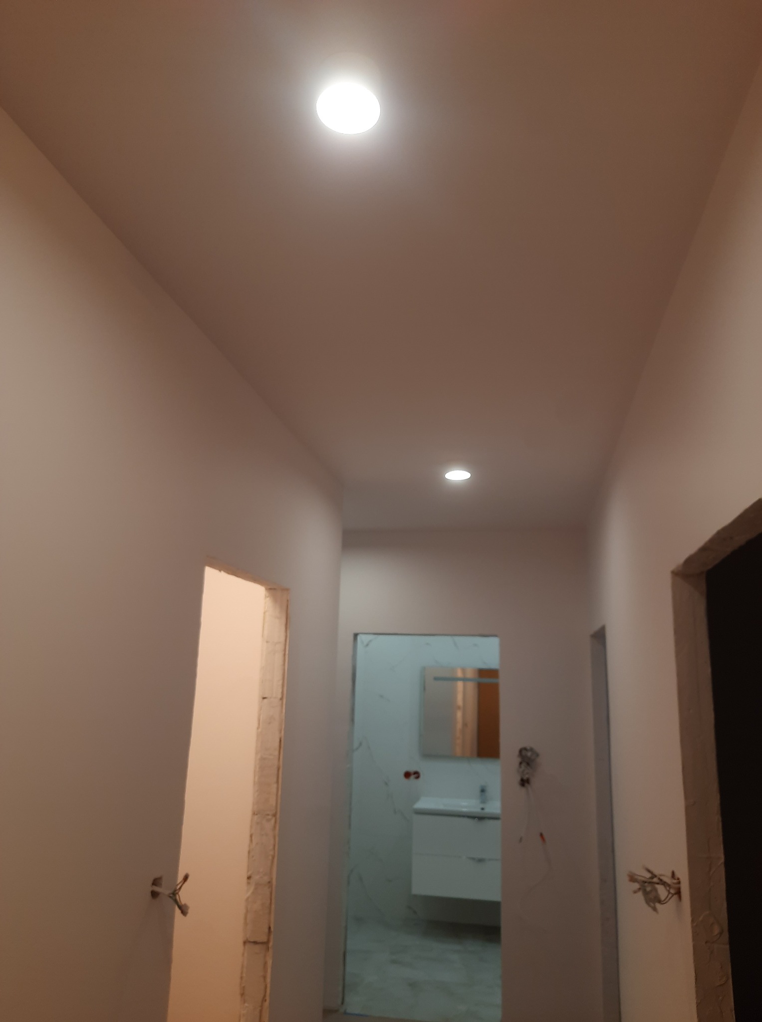 Korytarz po malowaniu, widoczne gładkie, jasne ściany i sufit z zamontowanymi lampami. W tle wykończona łazienka z umywalką i lustrem. Ościeżnice drzwiowe surowe.