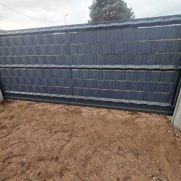 Usługi Spawalnicze - Przesuwna brama panelowa wypełniona tkaniną maskującą, osadzona pomiędzy betonowymi słupkami na żwirowym podłożu.