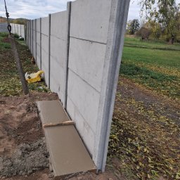 Usługi Spawalnicze - Montaż prefabrykowanego ogrodzenia betonowego: widoczny fragment wylewki fundamentu, pionowy panel ogrodzeniowy z widocznymi łączeniami, tło z zielenią i opadłymi liśćmi.