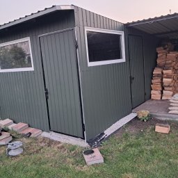 Usługi Spawalnicze - Zielona, blaszana konstrukcja z dwoma oknami i drzwiami, przylegająca do zadaszonej wiaty na drewno. Na pierwszym planie narzędzia spawalnicze i kostka brukowa.