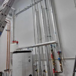 Usługi Spawalnicze - Biały zasobnik ciepłej wody użytkowej z podłączonymi rurami miedzianymi i stalowymi, drabina oparta o ścianę, instalacja w pomieszczeniu technicznym.