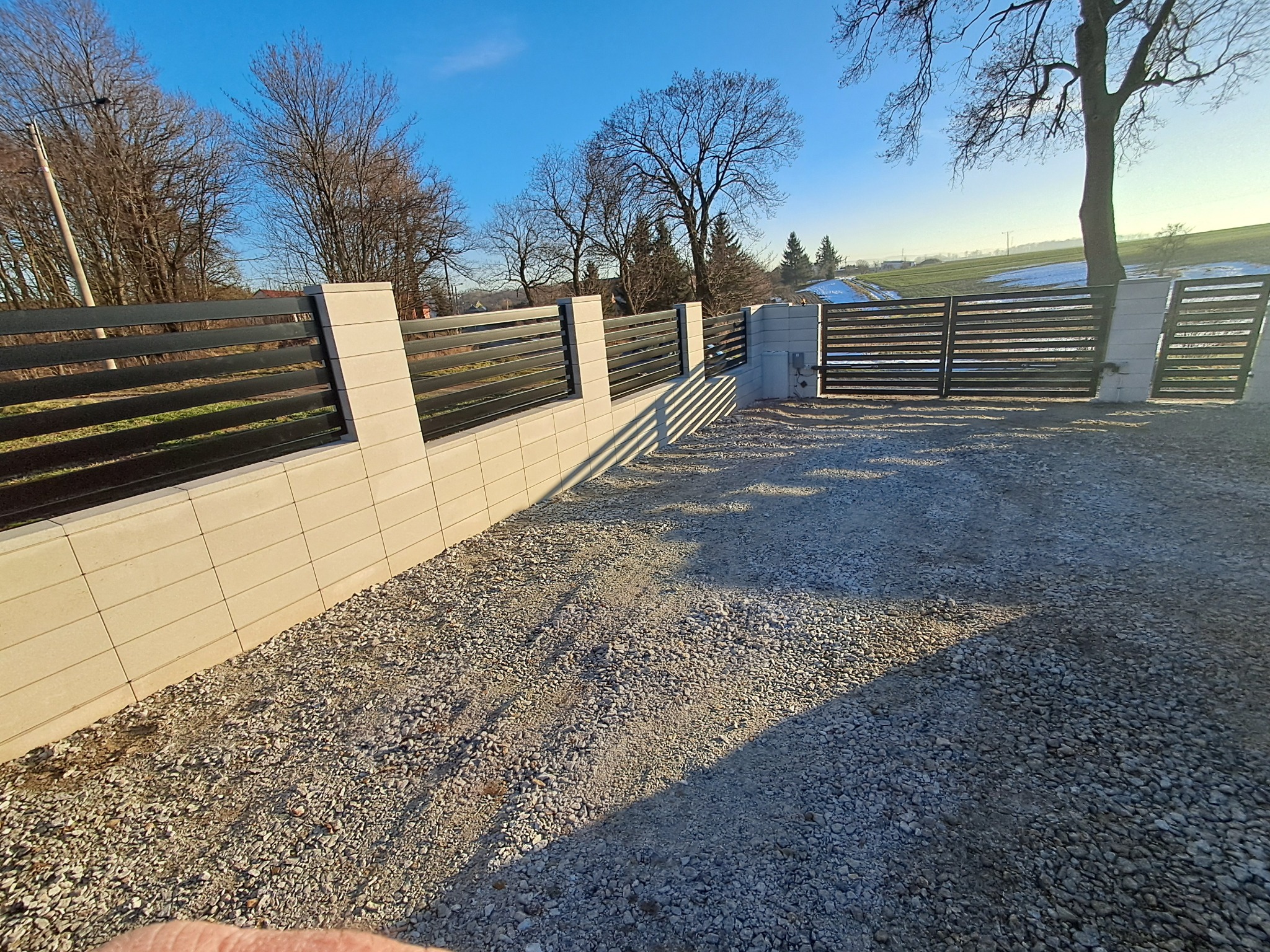 Nowoczesne ogrodzenie panelowe z poziomych, czarnych przęseł i jasnych, betonowych słupków na żwirowym podłożu, brama wjazdowa w tle, zimowy krajobraz z polami i drzewami.