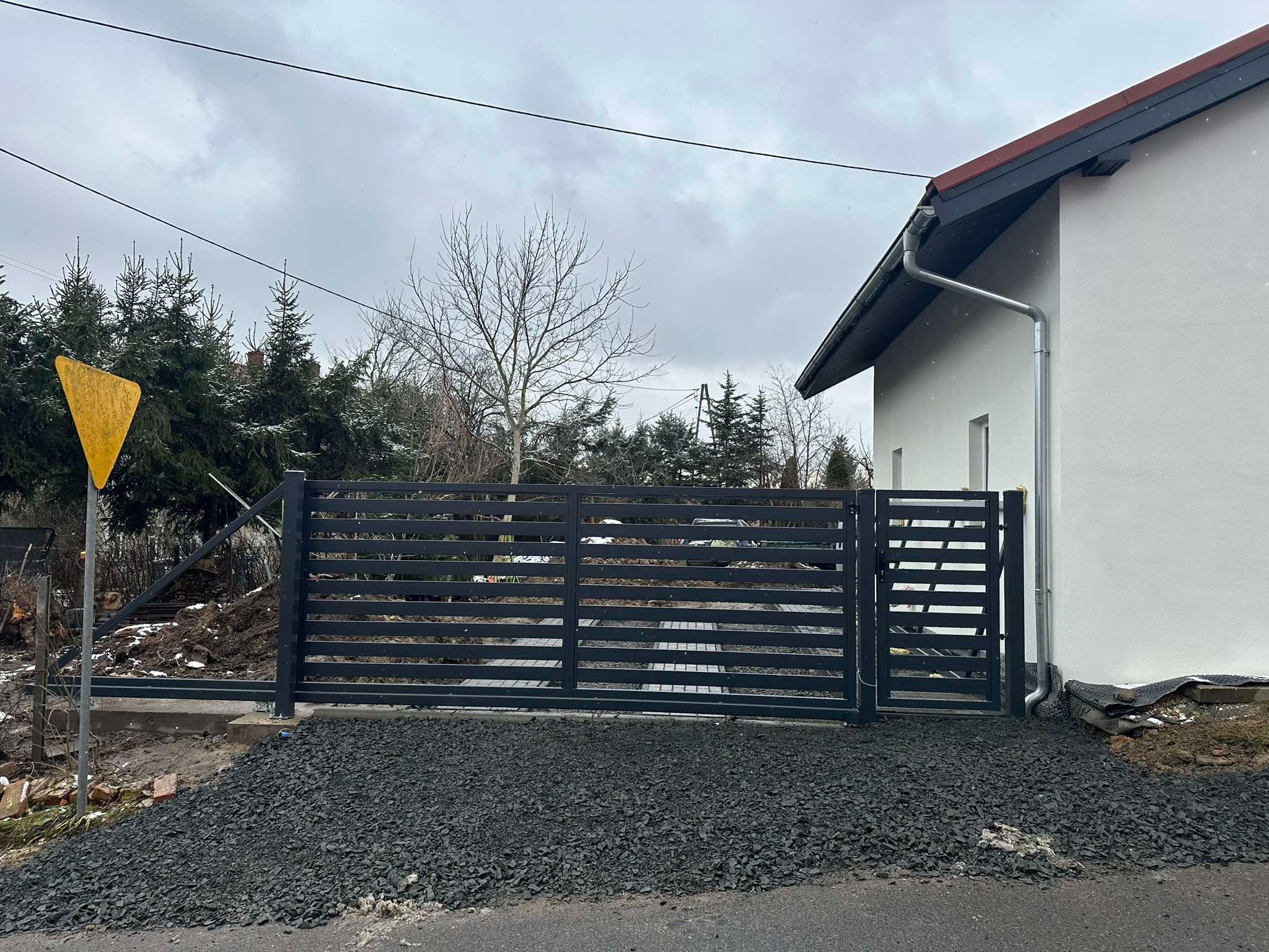 Nowoczesna, pozioma brama wjazdowa z metalowymi przęsłami w kolorze grafitowym, zamontowana przy nowym domu jednorodzinnym, widoczny fragment chodnika i czarne kamienie ozdobne