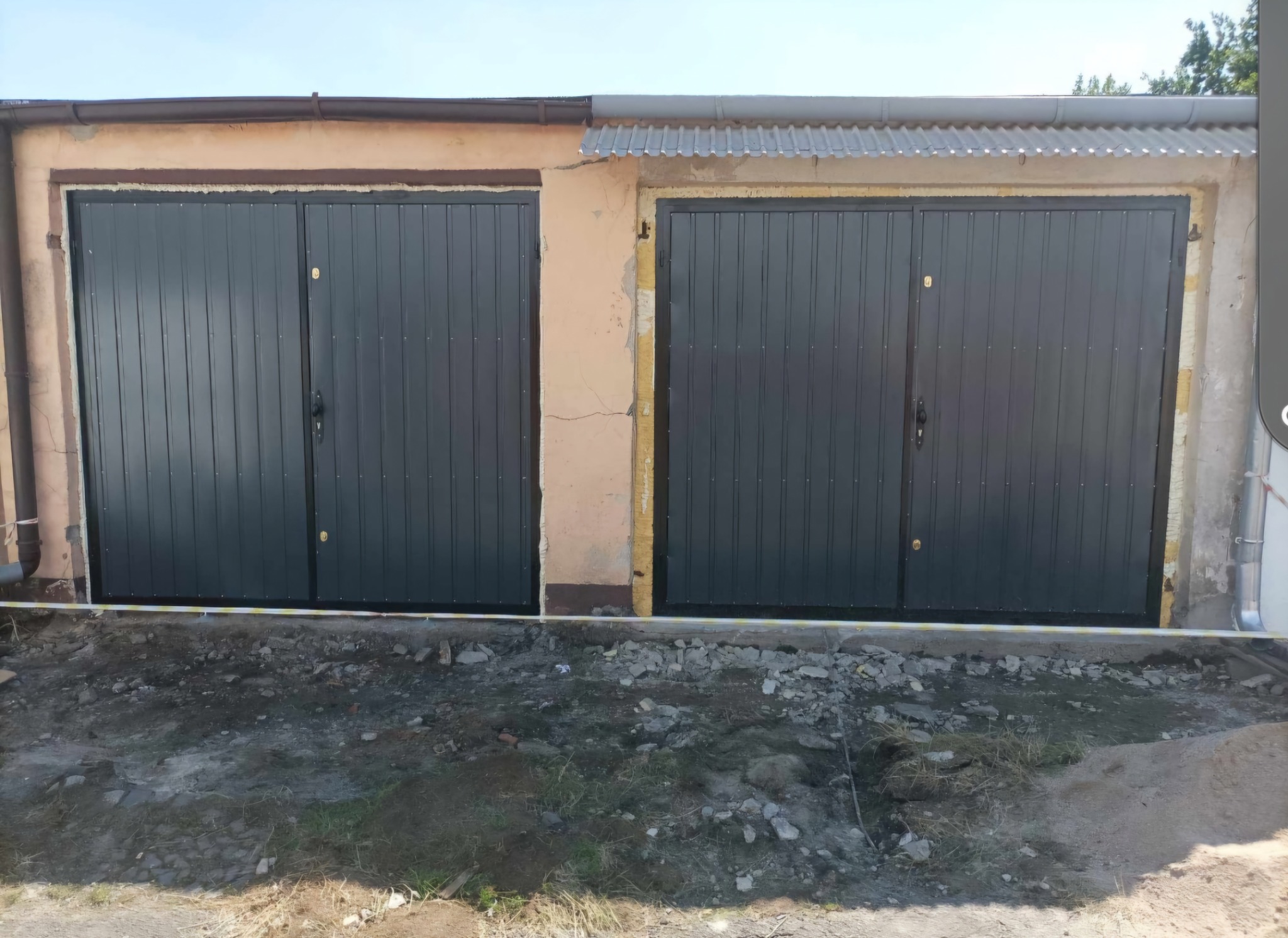 Dwa skrzydła grafitowej bramy garażowej z pionowymi panelami, zamontowane w budynku z beżową elewacją i fragmentem rynny, teren przed bramą w trakcie prac wykończeniowych.