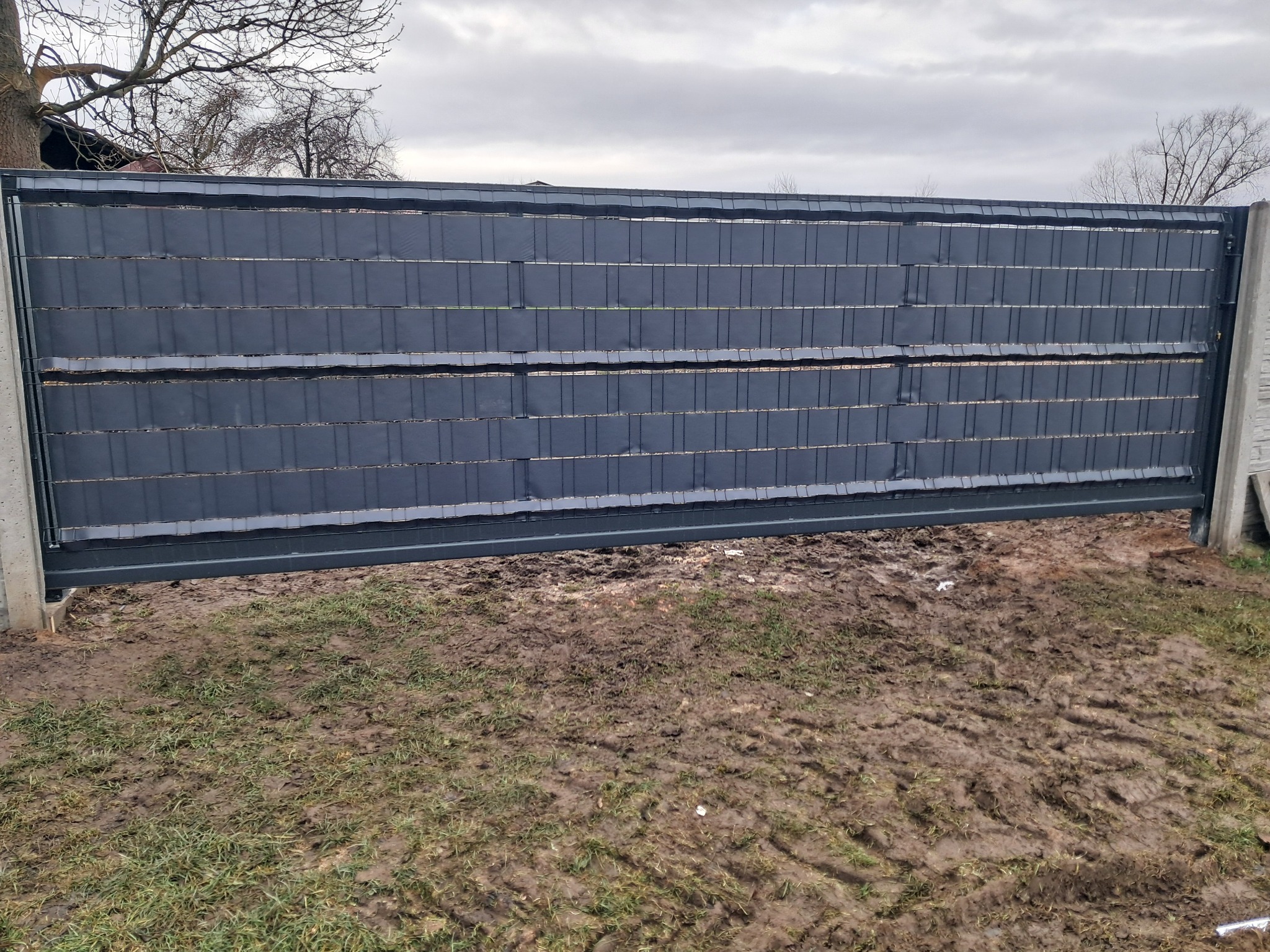 Przesuwna brama wjazdowa z poziomymi panelami kompozytowymi w kolorze grafitowym, zamontowana między betonowymi słupkami, na tle błotnistego trawnika i pochmurnego nieba.