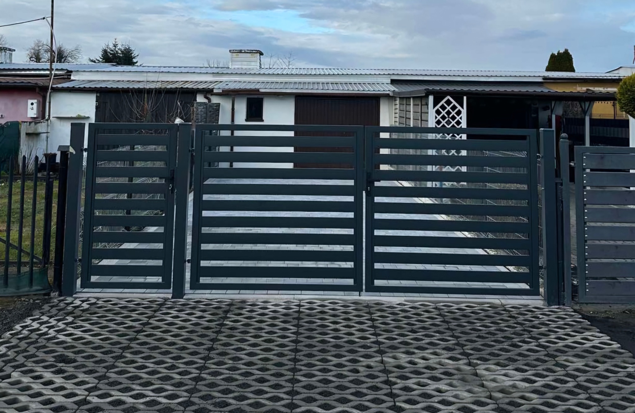 Nowoczesna, ciemnoszara brama wjazdowa z poziomymi panelami, ustawiona na podjeździe z ażurowych płyt betonowych. W tle widoczny dom z jasną elewacją i szarym dachem.