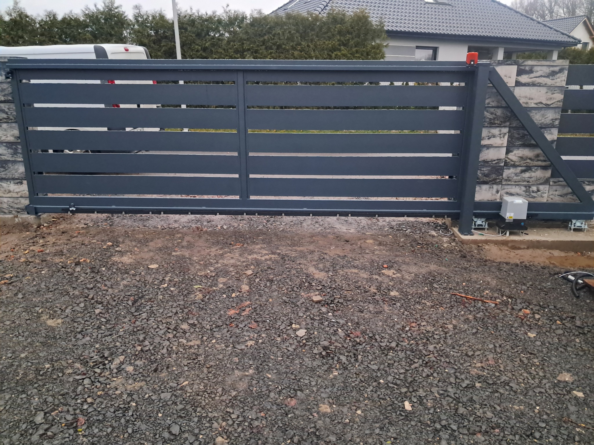 Szara przesuwna brama wjazdowa z poziomymi panelami, zamontowana na szarym kamiennym ogrodzeniu, widoczny automat do bramy, teren wokół wysypany żwirem.
