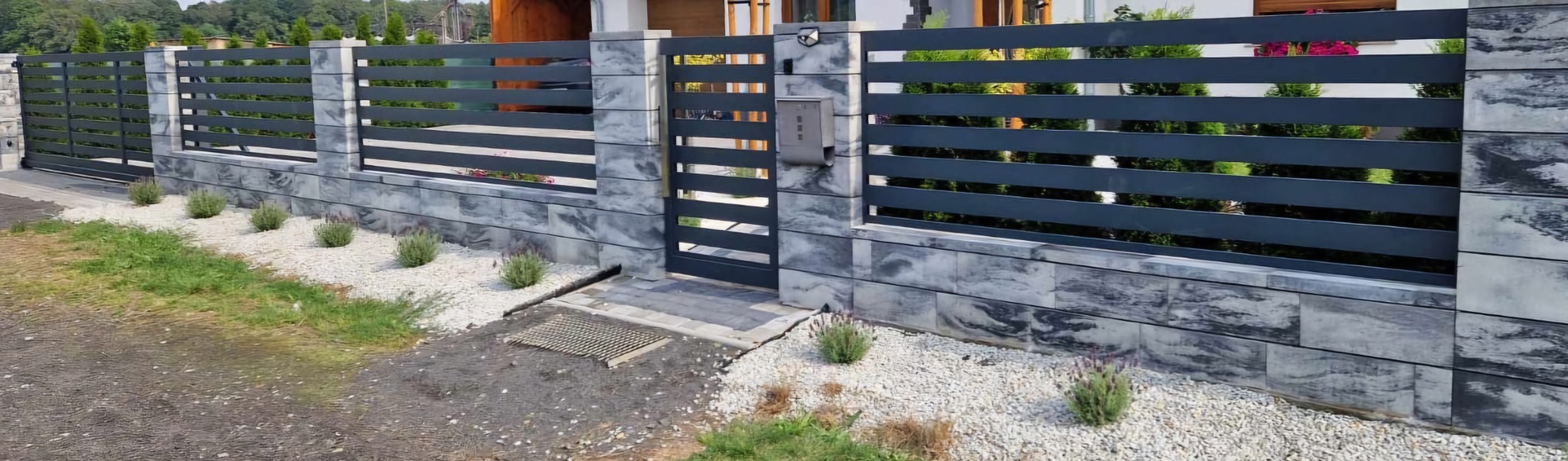 Nowoczesne ogrodzenie panelowe z poziomymi przęsłami w kolorze grafitowym, osadzone na murku z szarych bloczków imitujących kamień, z małym ogródkiem z białym grysem i nasadzeniami lawendy.