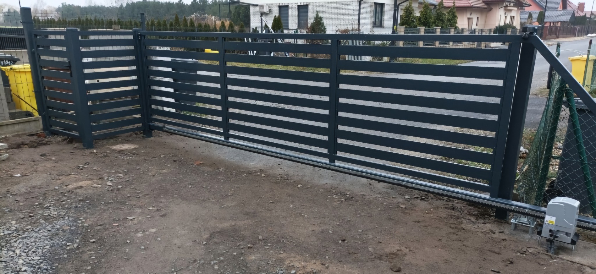 Nowoczesna, przesuwna brama wjazdowa z poziomymi panelami w kolorze grafitowym, widoczny automat do bramy i fragment ogrodzenia z siatki.