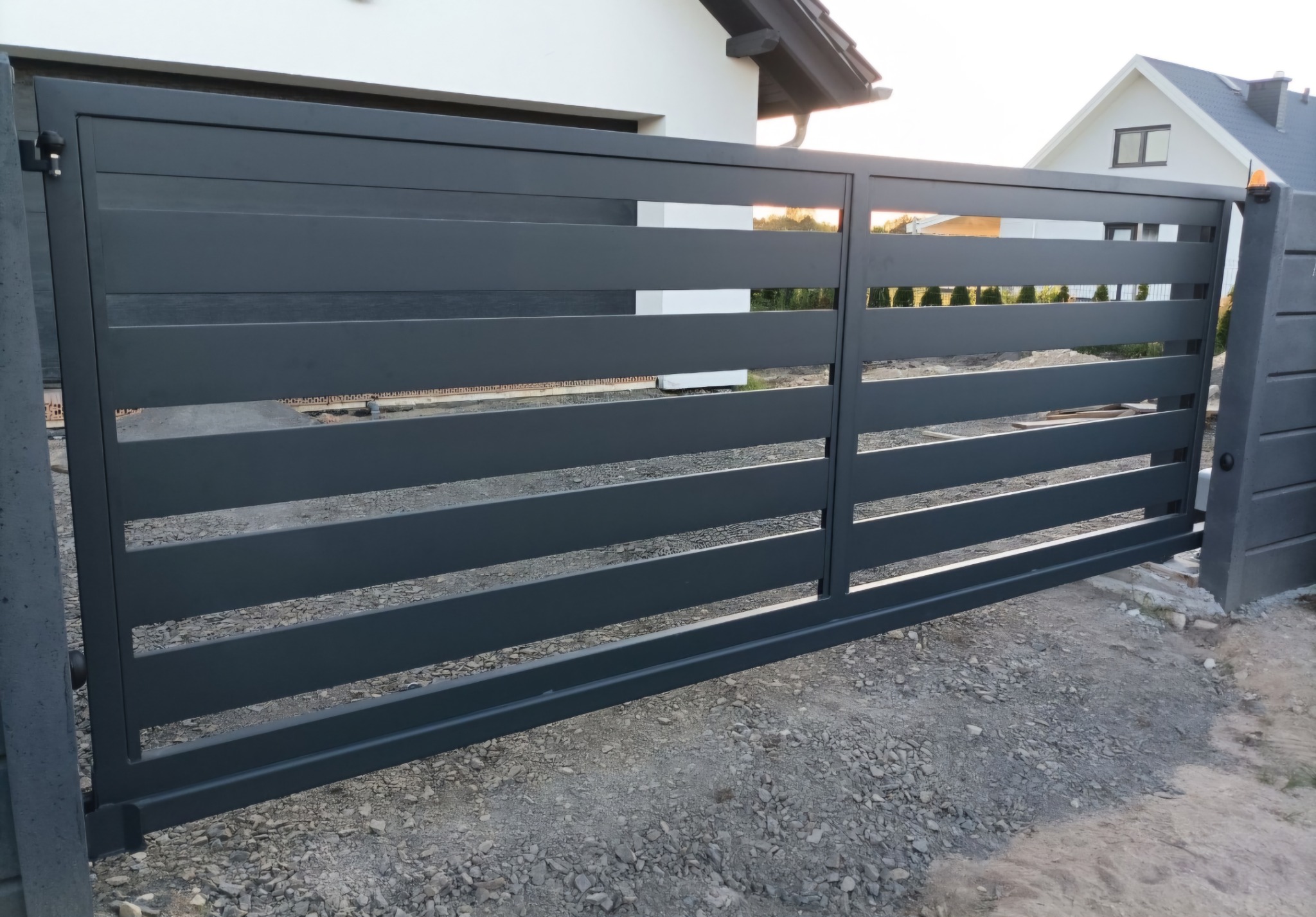 Nowoczesna, przesuwna brama wjazdowa w kolorze grafitowym z poziomymi profilami, na tle domu jednorodzinnego i żwirowego podłoża.