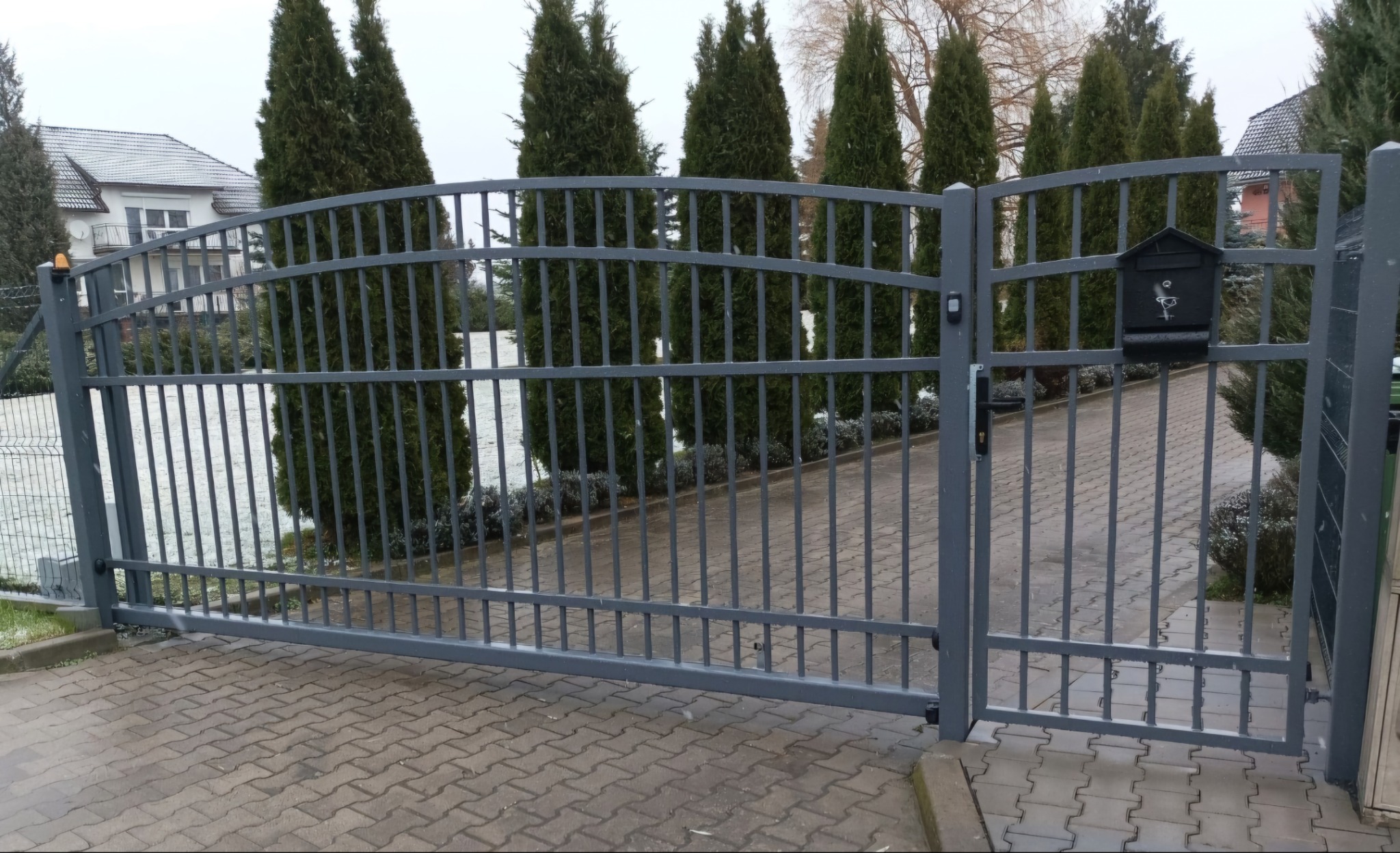 Szara brama wjazdowa z metalowymi prętami i łukowatym górnym panelem, skrzynka na listy z motywem ptaka po prawej stronie, widok na posesję z tujami i domem w tle.