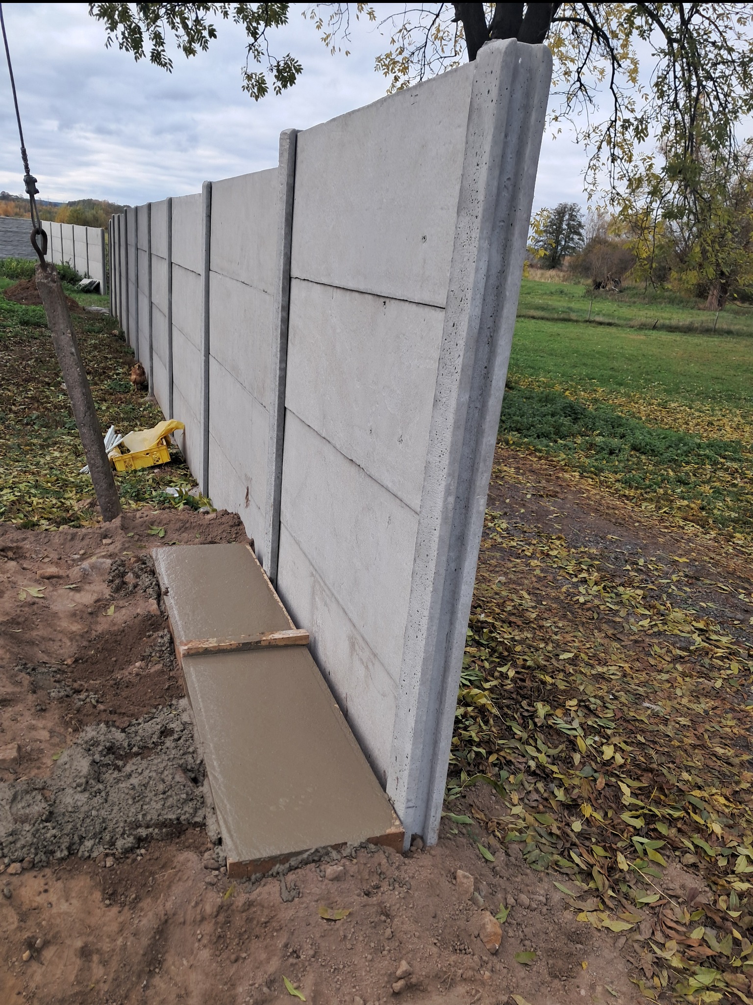 Montaż prefabrykowanego ogrodzenia betonowego: widoczny fragment wylewki fundamentu, pionowy panel ogrodzeniowy z widocznymi łączeniami, tło z zielenią i opadłymi liśćmi.