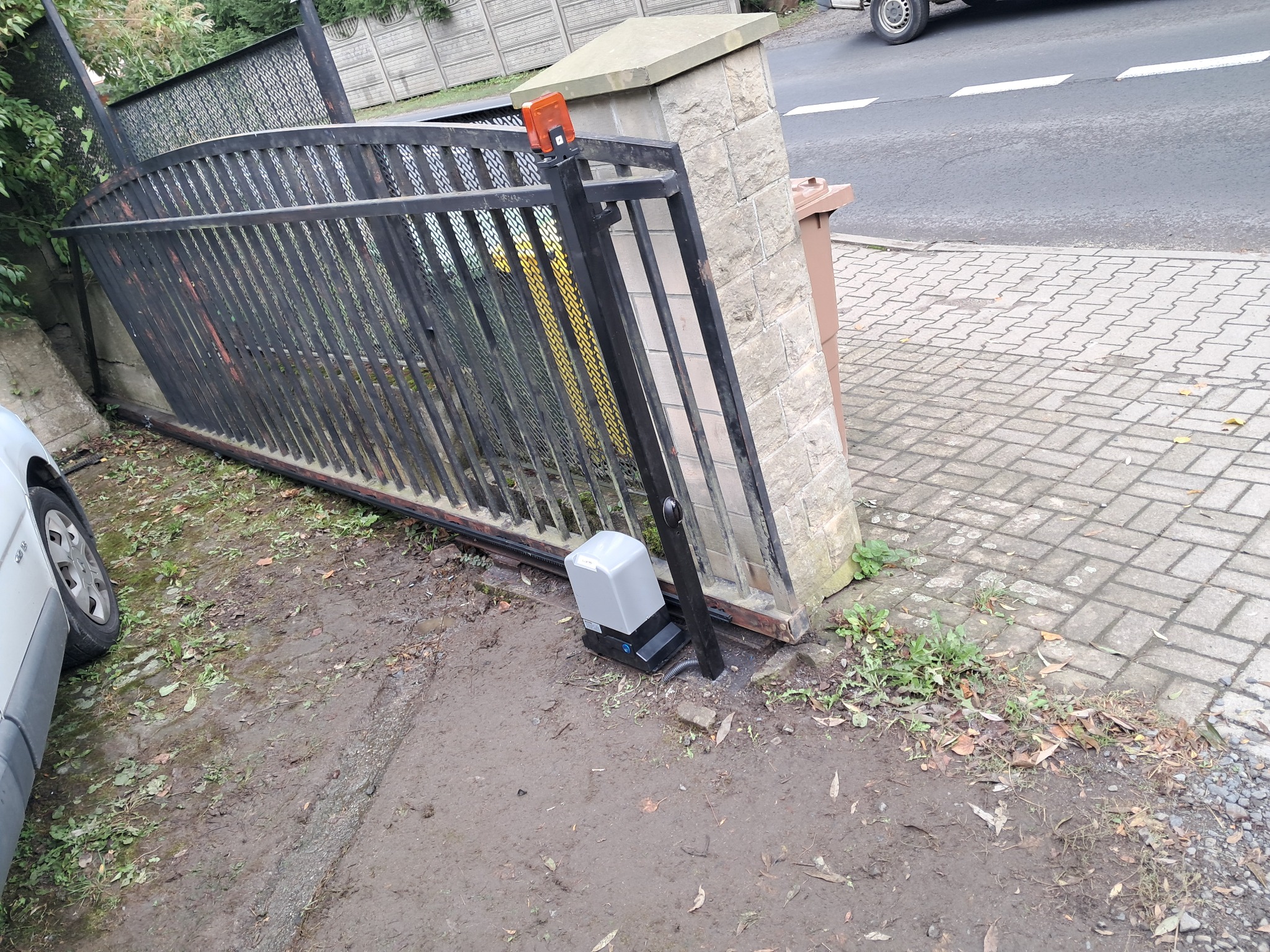 Czarna, metalowa brama wjazdowa z automatyką leżąca na ziemi obok słupka z kamienia, widoczny silnik napędu oraz fragment samochodu i chodnika.