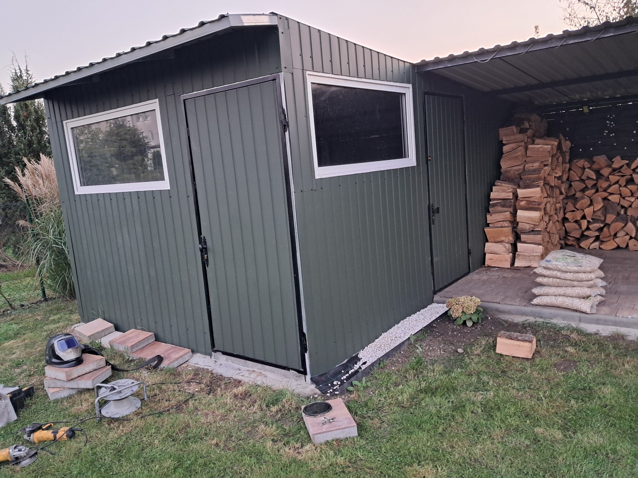 Zielona, blaszana konstrukcja z dwoma oknami i drzwiami, przylegająca do zadaszonej wiaty na drewno. Na pierwszym planie narzędzia spawalnicze i kostka brukowa.