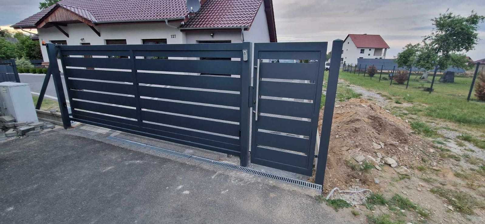 Nowoczesna, przesuwna brama wjazdowa z furtką w kolorze antracytowym, z poziomymi panelami, zamontowana przed domem z numerem 127, widoczny fragment odwodnienia liniowego.