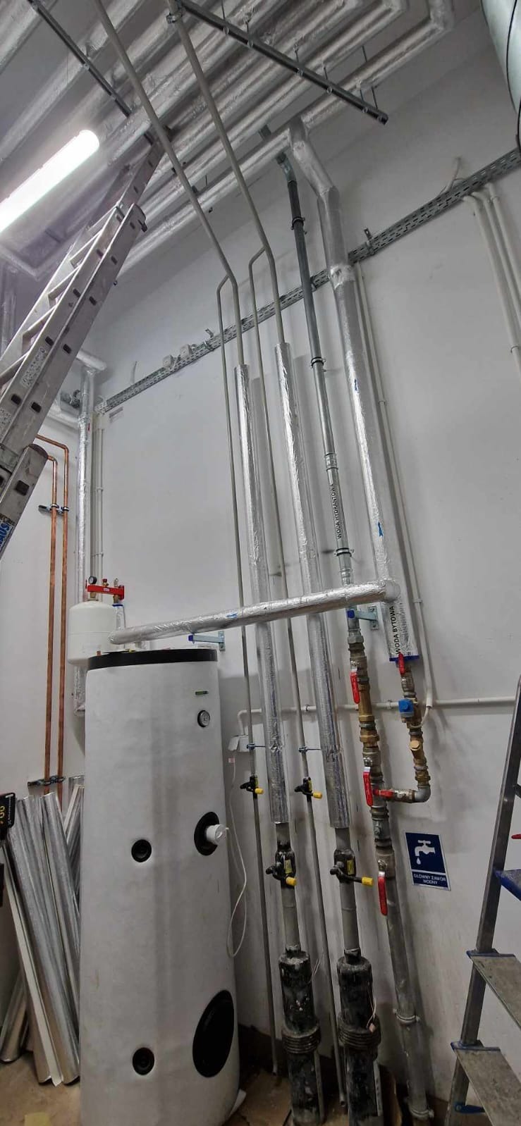 Biały zasobnik ciepłej wody użytkowej z podłączonymi rurami miedzianymi i stalowymi, drabina oparta o ścianę, instalacja w pomieszczeniu technicznym.