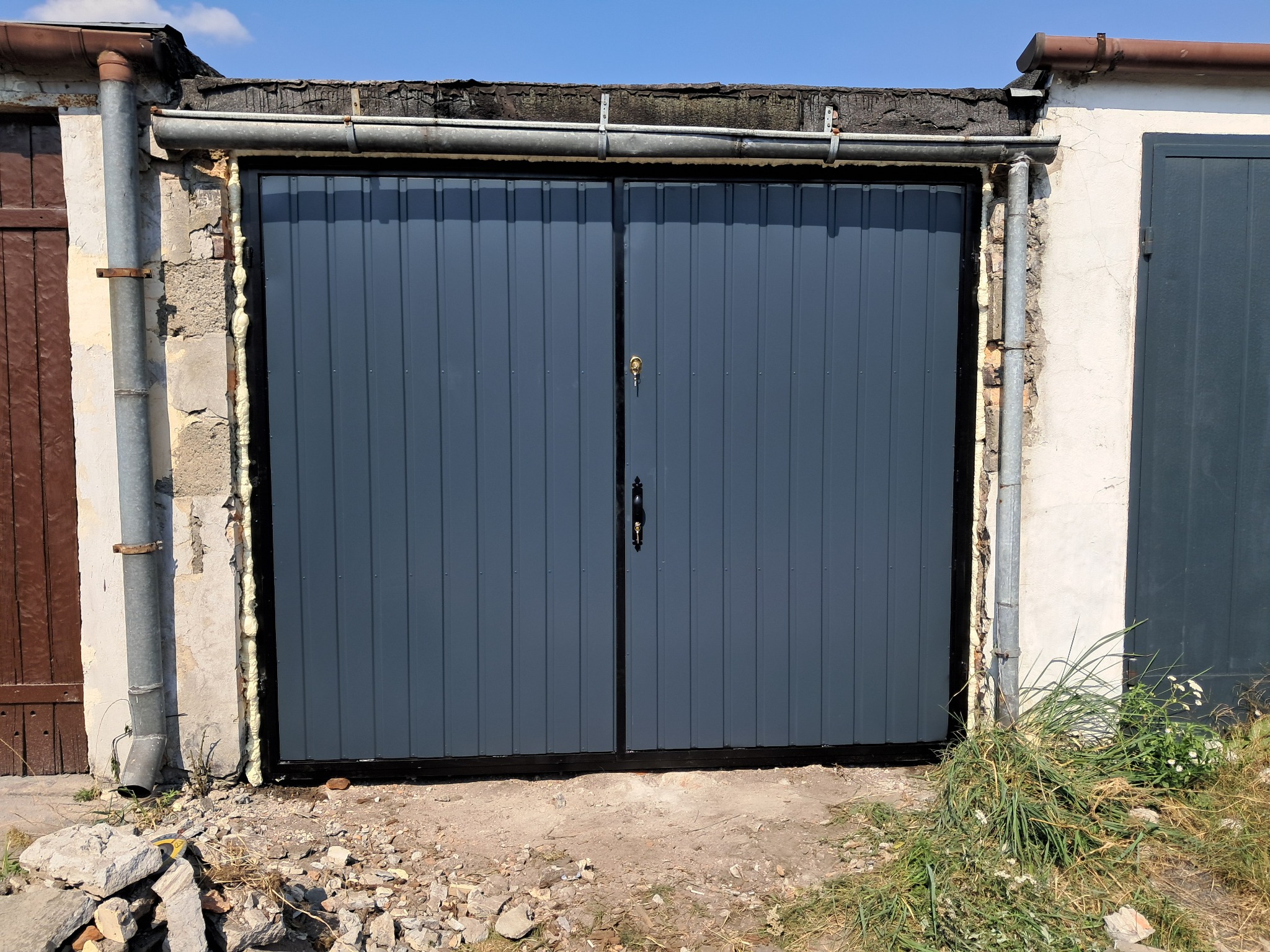 Szara, dwuskrzydłowa brama garażowa z widoczną pianką montażową wokół ościeżnicy, zamontowana w starej, częściowo zniszczonej ścianie budynku gospodarczego.