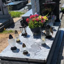 Granitowy nagrobek z kwiatami i zniczami na cmentarzu w słoneczny dzień. Czarny napis na płycie nagrobnej. Krzyż na pomniku. Widok na inne groby.