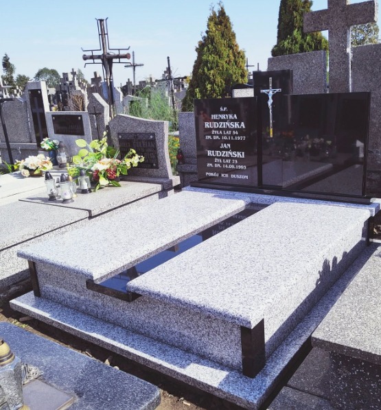 Granitowy nagrobek dwuosobowy z czarną tablicą inskrypcyjną i krzyżem na cmentarzu, w tle inne nagrobki i zieleń.