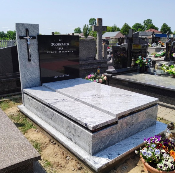 Jasnoszary nagrobek z czarną tablicą i krzyżem, na cmentarzu, w tle inne groby i zieleń. Widoczne kwiaty w doniczce. Nagrobek z jasnego kamienia.