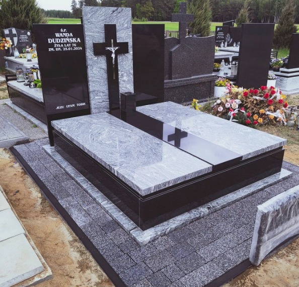 Granitowy nagrobek z krzyżem i inskrypcją 'Wanda Dudzińska', otoczony kwiatami na cmentarzu, w pochmurny dzień. Widok z bliska, skupienie na detalu.