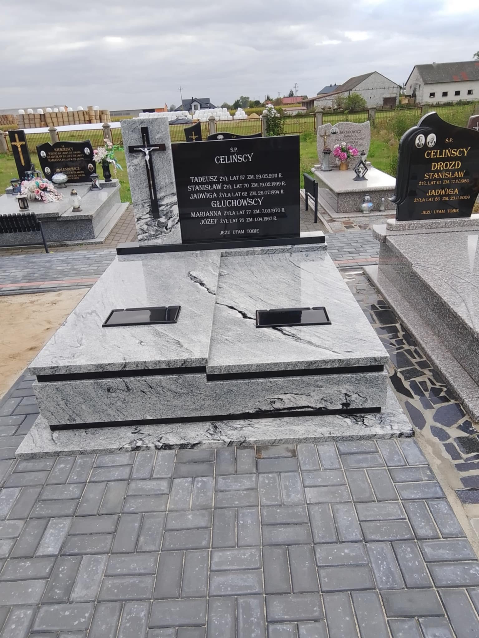 Nowoczesny nagrobek z jasnego kamienia o delikatnej fakturze, z czarną tablicą inskrypcyjną i krzyżem, na tle cmentarza z innymi pomnikami i zielenią.