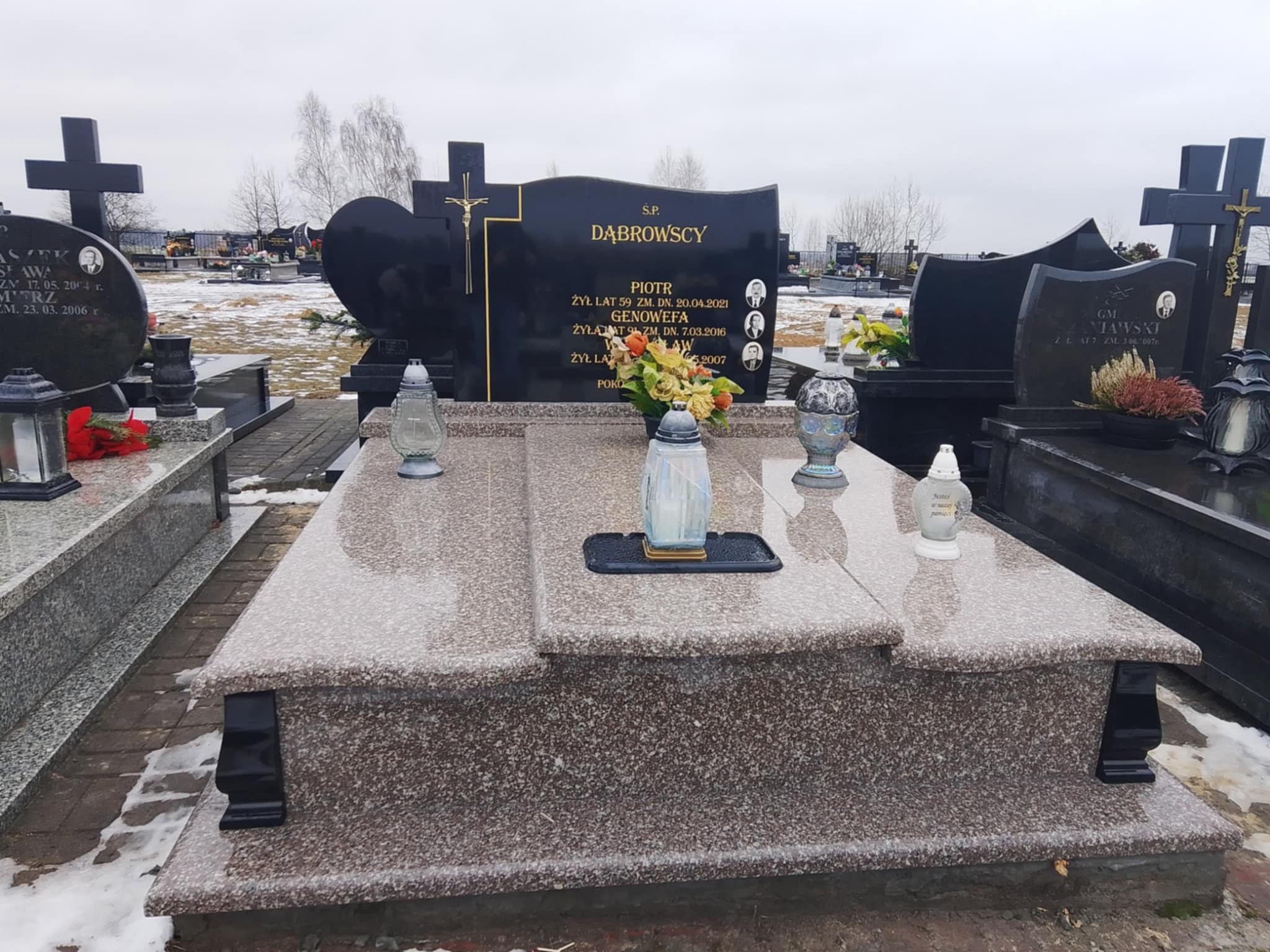 Granitowy nagrobek w odcieniach szarości i czerni, z wyrytymi danymi zmarłych i kwiatami. Na cmentarzu, w tle inne nagrobki i drzewa bez liści.