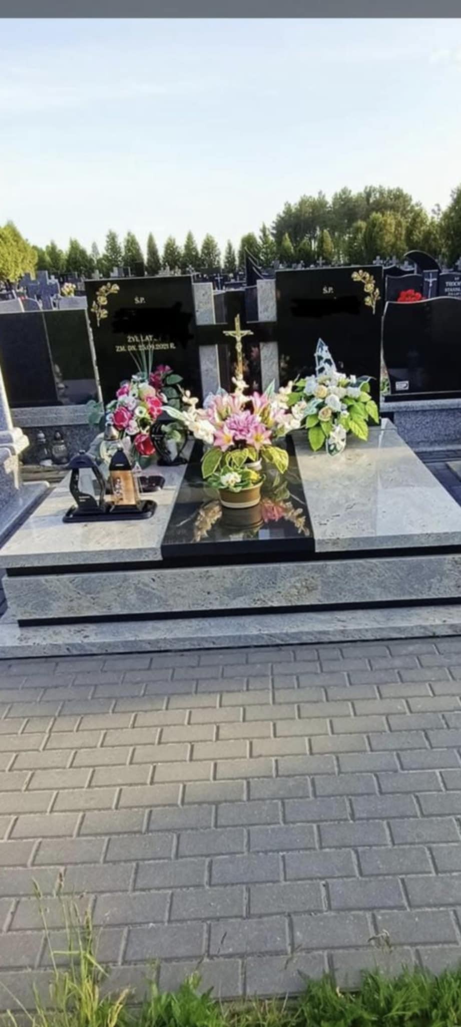 Podwójny, granitowy nagrobek z czarnymi płytami, zdobiony kwiatami i krzyżem, na cmentarzu. Widoczna kostka brukowa i drzewa w tle.