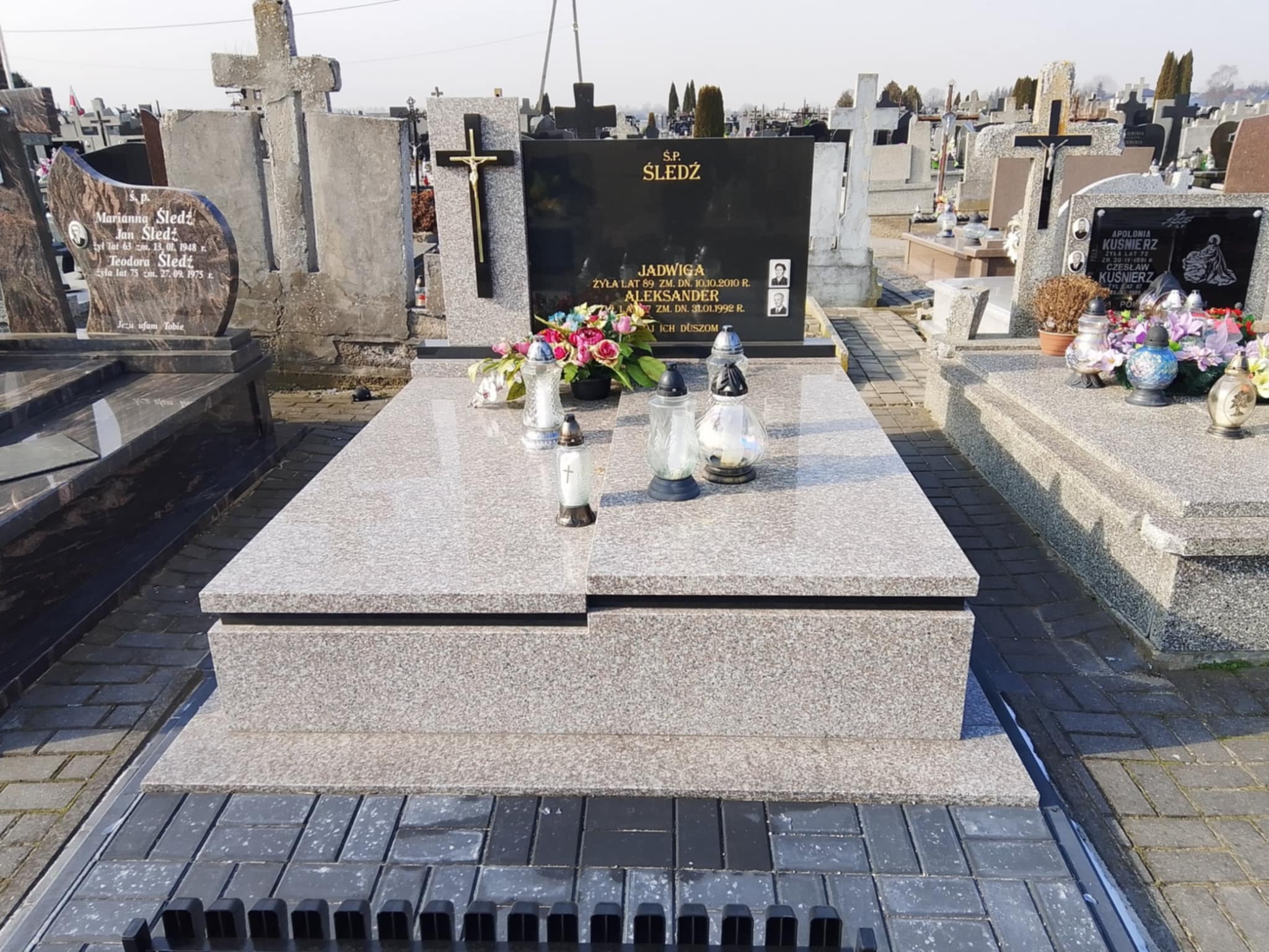 Granitowy nagrobek z czarną tablicą, zdobiony kwiatami i zniczami, na cmentarzu z licznymi grobami w tle. Uwagę przykuwa jasny odcień kamienia.