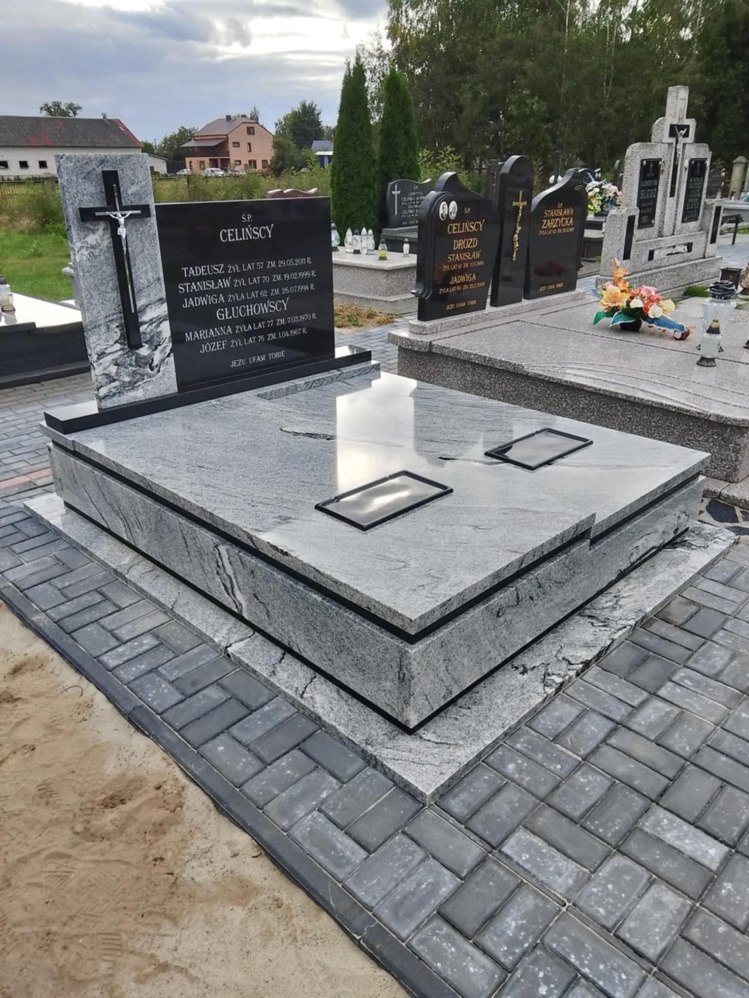 Szary, granitowy nagrobek z czarną tablicą inskrypcyjną i krzyżem, otoczony kostką brukową na cmentarzu. Widoczne inne nagrobki i zieleń w tle.