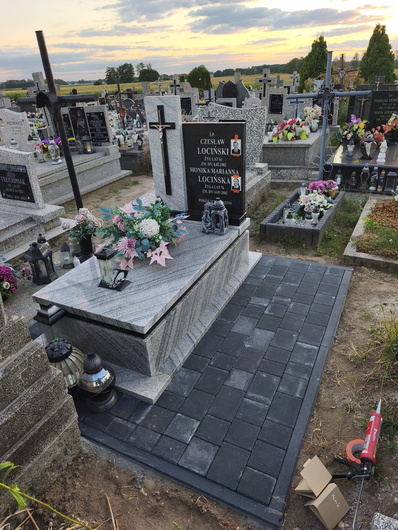 Granitowy nagrobek z czarną tablicą, ozdobiony kwiatami i lampionami, na cmentarzu. Ułożona kostka brukowa wokół pomnika. Widoczne narzędzia montażowe.