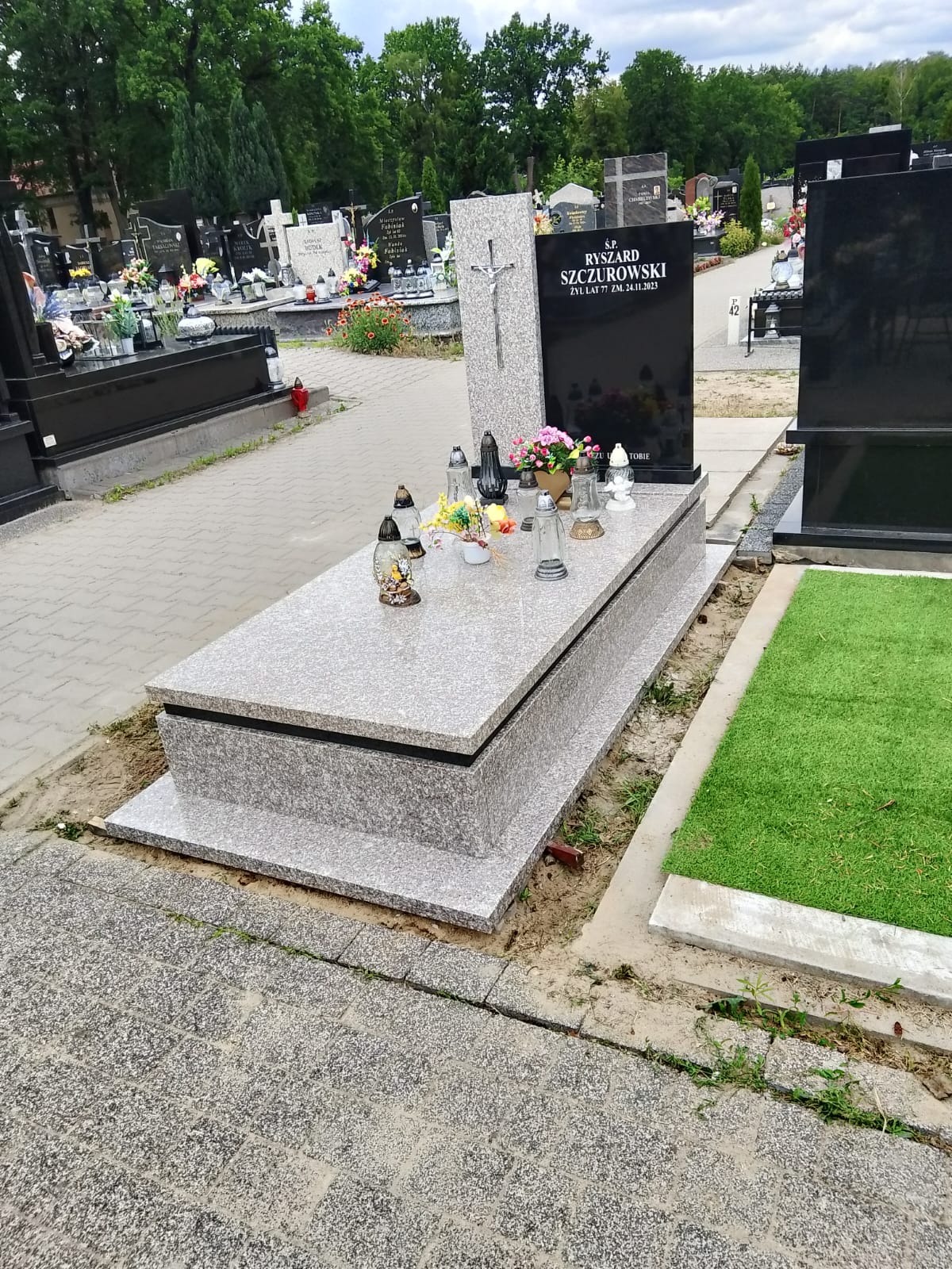 Jasnoszary nagrobek z czarną tablicą i krzyżem, otoczony kwiatami i zniczami na cmentarzu, z kostką brukową i fragmentem sztucznej trawy. Widok z boku.