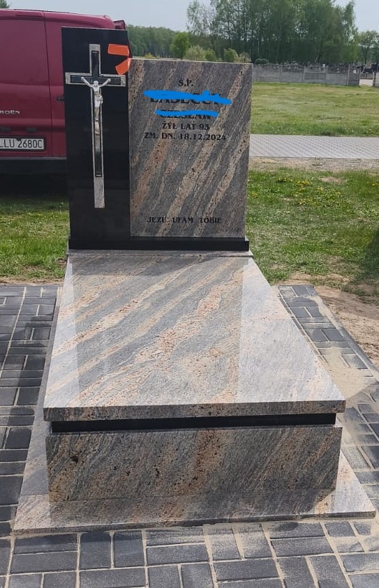 Granitowy nagrobek z czarnym elementem i srebrnym krzyżem, umieszczony na cmentarzu. Widoczny fragment czerwonego samochodu z boku. Napis: Jezu ufam Tobie.