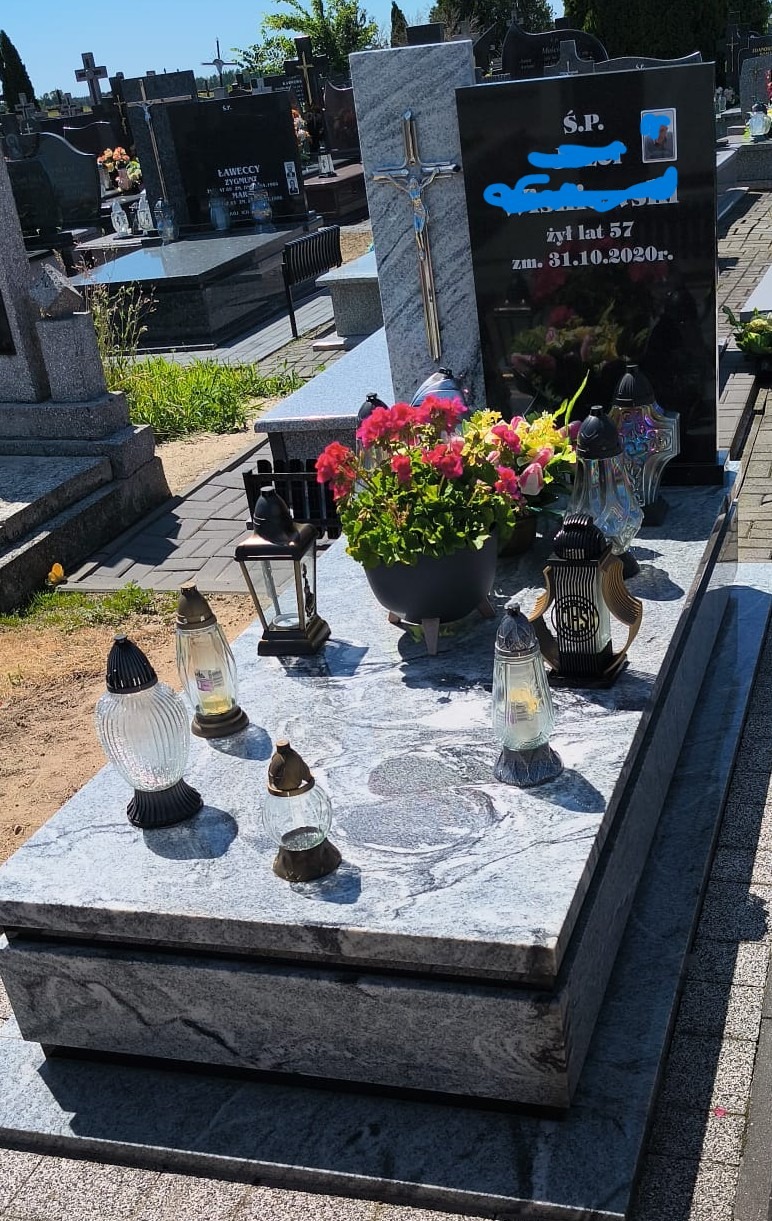 Granitowy nagrobek z kwiatami i zniczami na cmentarzu w słoneczny dzień. Czarny napis na płycie nagrobnej. Krzyż na pomniku. Widok na inne groby.