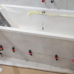 Biała wanna zabezpieczona folią podczas układania płytek imitujących beton na obudowie, z użyciem poziomicy i klipsów poziomujących.