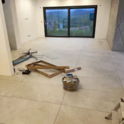 Układanie beżowych płytek podłogowych w jasnym pokoju z dużym oknem; widoczne narzędzia glazurnicze, wiadro z zaprawą i drabina.