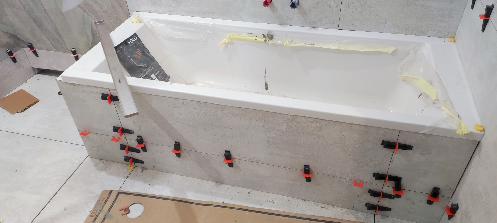 Biała wanna zabezpieczona folią podczas układania płytek imitujących beton na obudowie, z użyciem poziomicy i klipsów poziomujących.