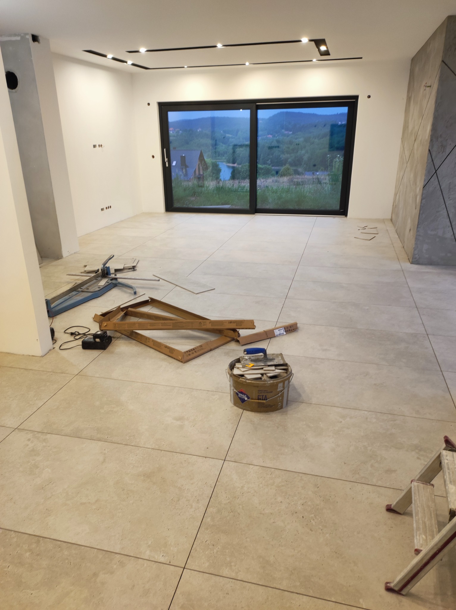 Układanie beżowych płytek podłogowych w jasnym pokoju z dużym oknem; widoczne narzędzia glazurnicze, wiadro z zaprawą i drabina.