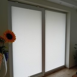 GREEN HOUSE Plejada-Stary Nowy Dom - Białe plisy okienne zamontowane na dwóch oknach, widok z wnętrza, z boku dekoracyjny bukiet sztucznych kwiatów.