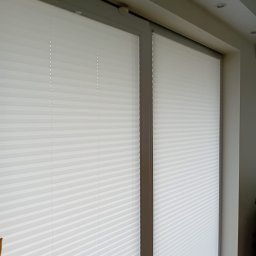 GREEN HOUSE Plejada-Stary Nowy Dom - Dwa okna z zamontowanymi białymi plisami, widoczne mechanizmy sterujące, jasne wnętrze z oświetleniem sufitowym.